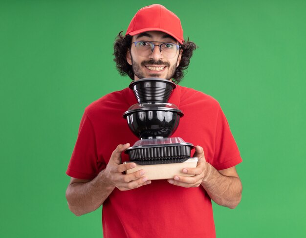 Repartidor joven sonriente en uniforme rojo y gorra con gafas sosteniendo paquetes de comida de papel y recipientes de comida debajo de la barbilla mirando al frente aislado en la pared verde