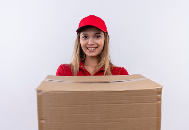 Repartidor joven sonriente con uniforme rojo y gorra sosteniendo una caja grande aislada en la pared blanca