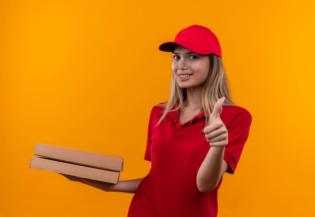 Repartidor joven sonriente con uniforme rojo y gorra sosteniendo la caja de pizza con el pulgar hacia arriba aislado en la pared naranja