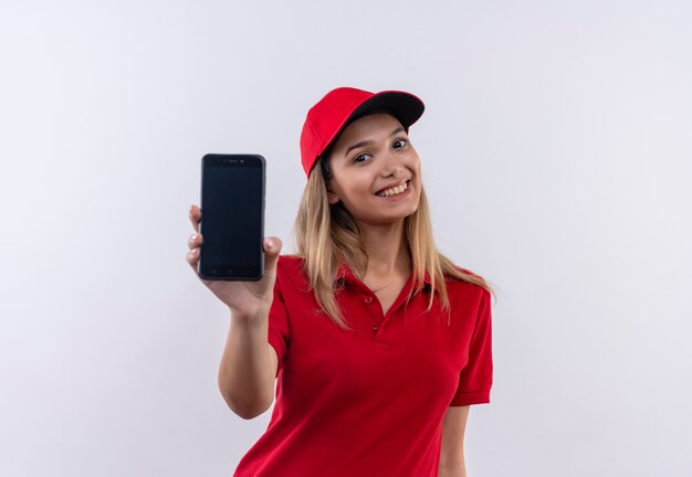 Repartidor joven sonriente con uniforme rojo y gorra sosteniendo el teléfono a la cámara aislada en blanco