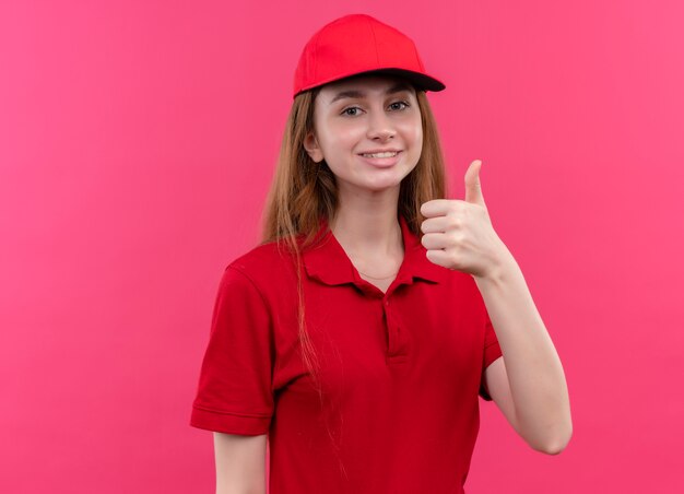 Repartidor joven sonriente en uniforme rojo que muestra el pulgar hacia arriba en la pared rosa aislada con espacio de copia