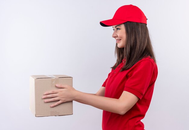 Repartidor joven sonriente vistiendo camiseta roja con gorra roja sosteniendo una caja en la pared blanca aislada
