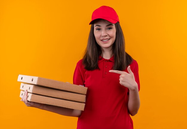 Repartidor joven sonriente vistiendo camiseta roja con gorra roja sosteniendo una caja de pizza y puntos con un dedo en la pared naranja aislada