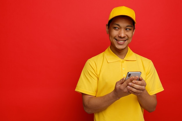 Repartidor joven sonriente vistiendo gorra y uniforme sosteniendo teléfono móvil mirando al lado