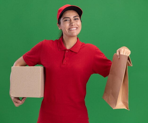 Repartidor joven sonriente vistiendo uniforme y caja de sujeción de gorra con paquete de alimentos de papel aislado en la pared verde