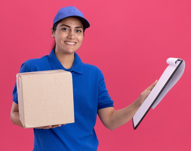 Repartidor joven sonriente vistiendo uniforme con caja de sujeción de tapa con portapapeles aislado en pared rosa