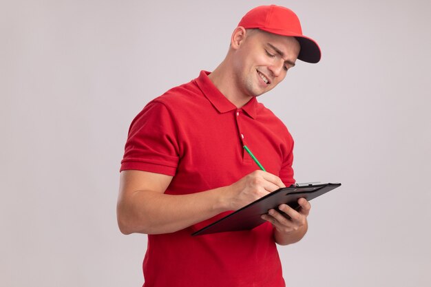 Repartidor joven sonriente vistiendo uniforme con gorra escribiendo algo en el portapapeles aislado en la pared blanca