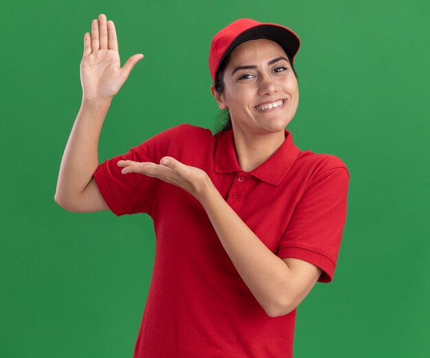 Repartidor joven sonriente vistiendo uniforme y gorra fingiendo sostener algo aislado en la pared verde