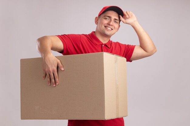Repartidor joven sonriente vistiendo uniforme con gorra sosteniendo una caja grande y gorra aislada en la pared blanca