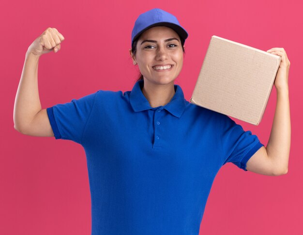 Repartidor joven sonriente vistiendo uniforme con gorra sosteniendo la caja en el hombro mostrando un gesto fuerte aislado en la pared rosa