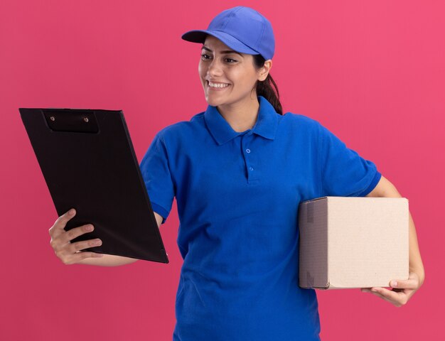 Repartidor joven sonriente vistiendo uniforme con gorra sosteniendo la caja y mirando el portapapeles en su mano aislada en la pared rosa