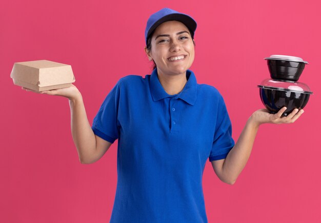 Repartidor joven sonriente vistiendo uniforme con gorra sosteniendo contenedores de alimentos aislados en la pared rosa