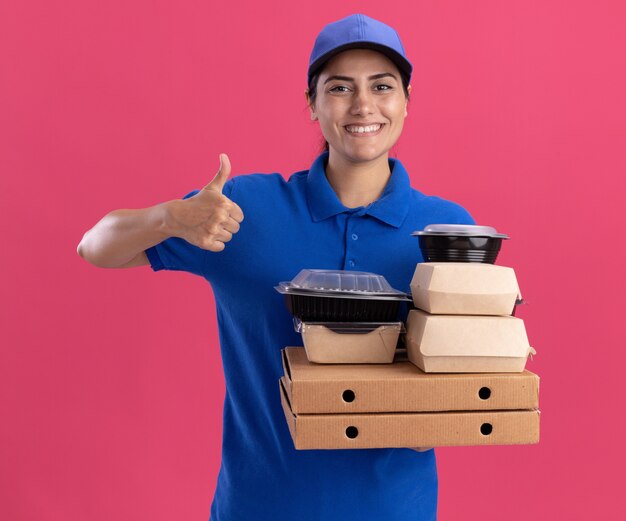 Repartidor joven sonriente vistiendo uniforme con gorra sosteniendo contenedores de comida en cajas de pizza mostrando el pulgar hacia arriba aislado en la pared rosa