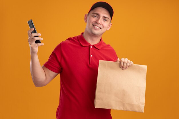 Repartidor joven sonriente vistiendo uniforme con gorra sosteniendo el paquete de alimentos de papel con teléfono aislado en la pared naranja