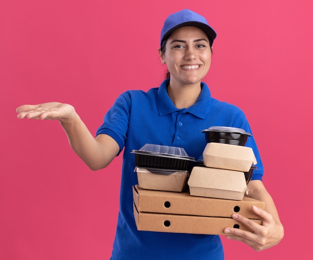 Repartidor joven sonriente vistiendo uniforme con tapa sosteniendo contenedores de comida en puntos de cajas de pizza con la mano al lado aislado en la pared rosa