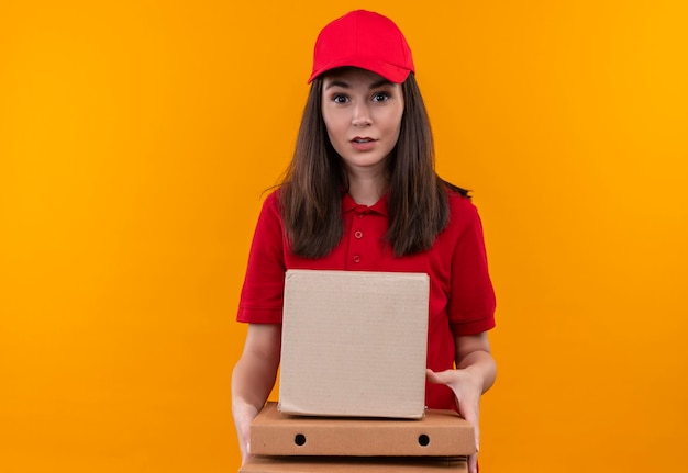 Foto gratuita repartidor joven sorprendido con camiseta roja con gorra roja sosteniendo una caja y caja de pizza en la pared amarilla aislada