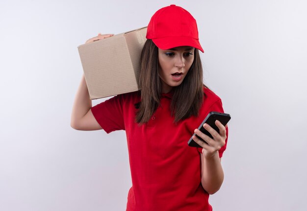 Repartidor joven sorprendido con camiseta roja con gorra roja, un teléfono y una caja en su hombro en una pared blanca aislada