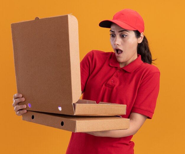 Repartidor joven sorprendido con uniforme y gorra sosteniendo y abriendo la caja de pizza aislada en la pared naranja