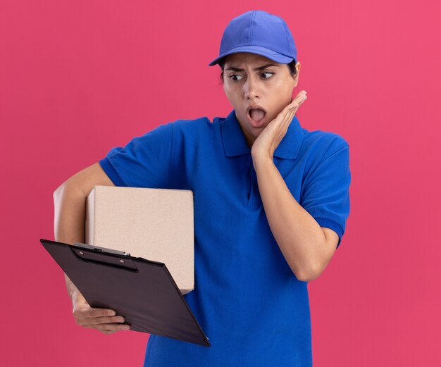 Repartidor joven sorprendido con uniforme con gorra sosteniendo la caja y mirando el portapapeles en la mano poniendo la mano en la mejilla aislada en la pared rosa