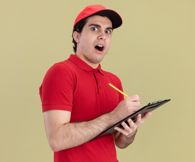 Repartidor joven sorprendido en uniforme rojo y gorra sosteniendo lápiz y portapapeles mirando al frente aislado en la pared verde oliva