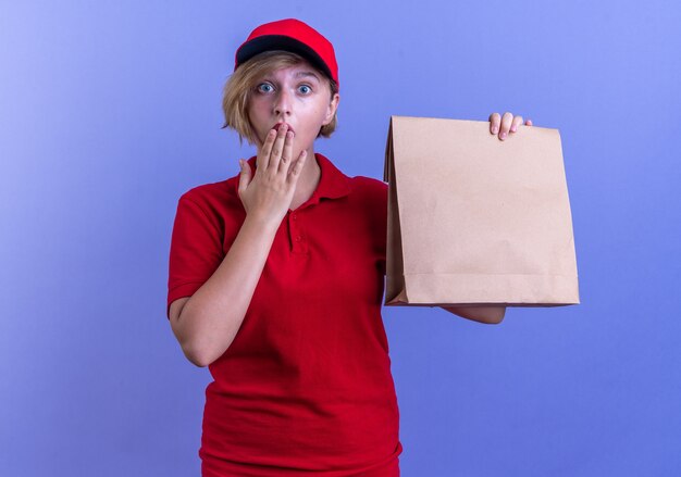 Repartidor joven sorprendido vistiendo uniforme y gorra sosteniendo una bolsa de comida de papel con la boca cubierta con la mano aislada en la pared azul