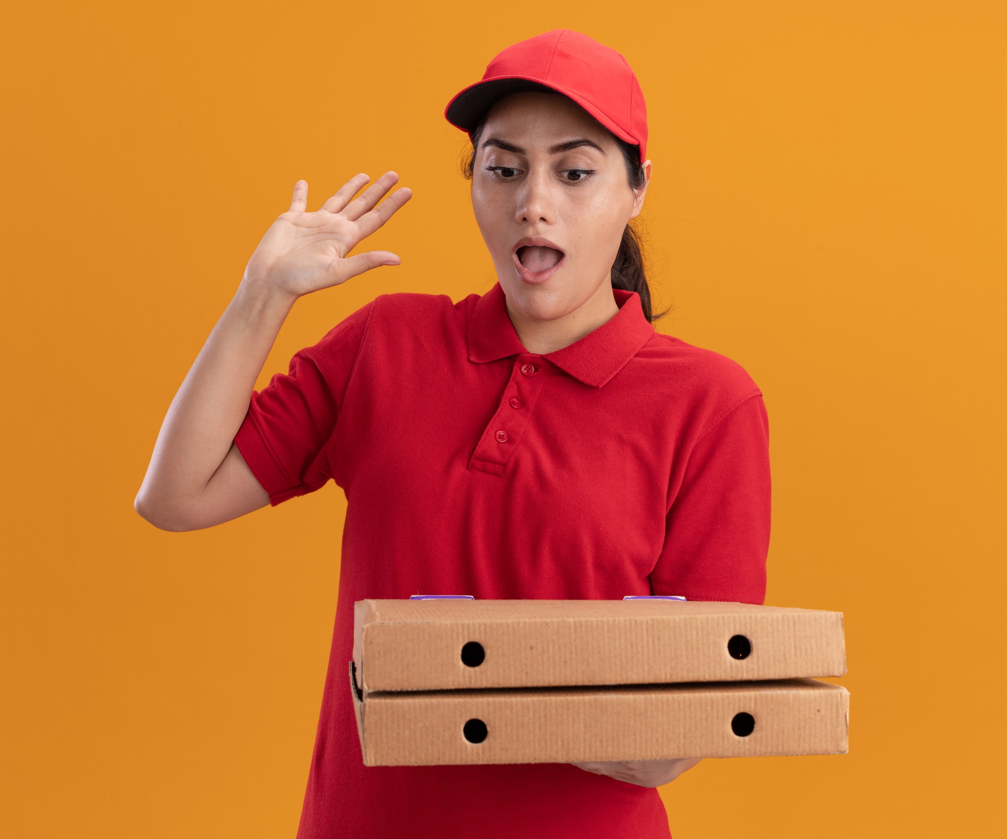 Repartidor joven sorprendido vistiendo uniforme y gorra sosteniendo y mirando cajas de pizza levantando la mano aislada en la pared naranja