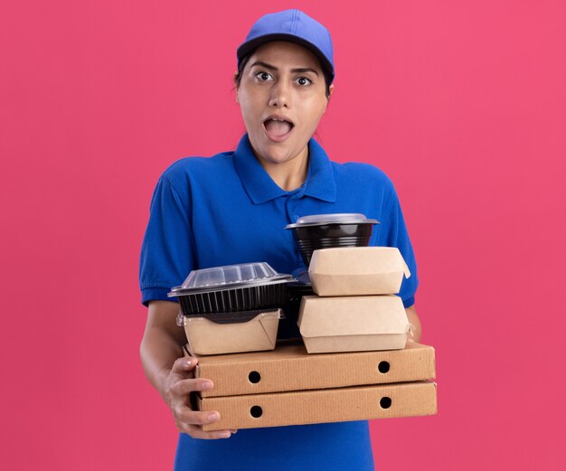 Repartidor joven sorprendido vistiendo uniforme con tapa sosteniendo contenedores de comida en cajas de pizza aisladas en la pared rosa