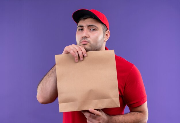 Repartidor joven tímido en uniforme rojo y gorra sosteniendo el paquete de papel confundido parado sobre la pared púrpura