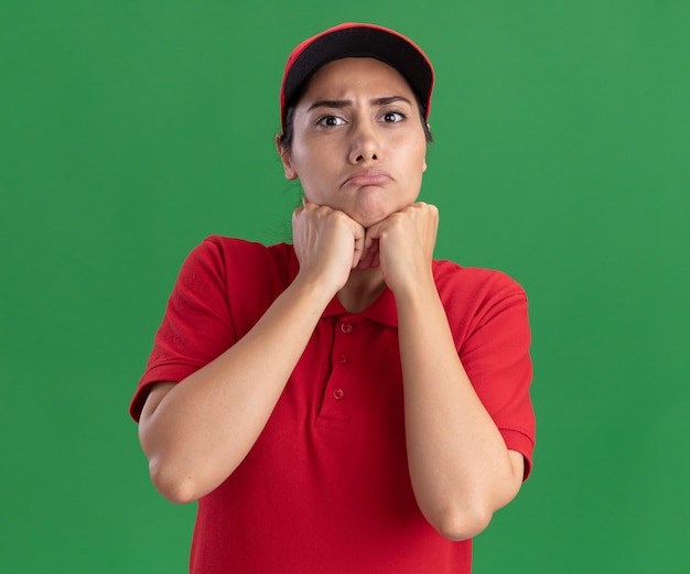 Repartidor joven triste vistiendo uniforme y gorra poniendo puños debajo de la barbilla aislado en la pared verde