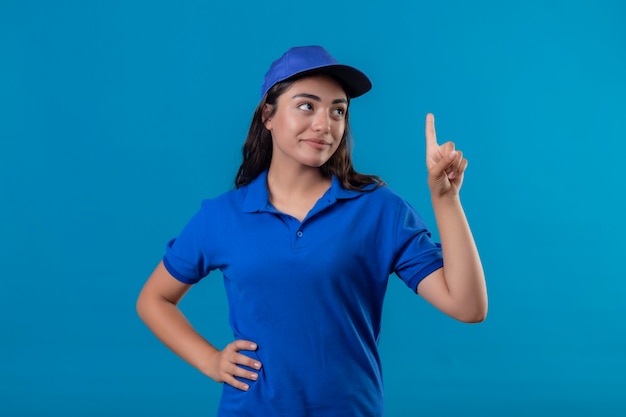 Repartidor joven en uniforme azul y gorra apuntando con el dedo hacia arriba pensando en positivo con sonrisa de confianza en la cara de pie sobre fondo azul.