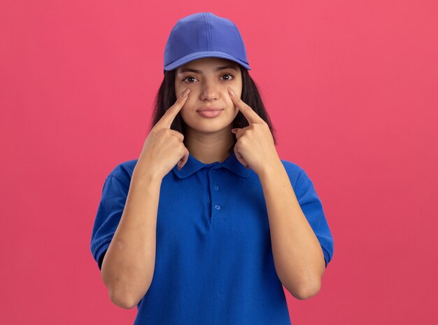 Repartidor joven en uniforme azul y gorra apuntando con el dedo índice a los ojos de pie sobre la pared rosa