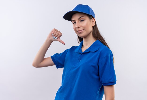 Repartidor joven en uniforme azul y gorra apuntando a sí misma, satisfecha y orgullosa