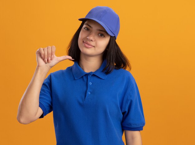 Repartidor joven en uniforme azul y gorra apuntando a sí misma sonriendo confiada de pie sobre la pared naranja