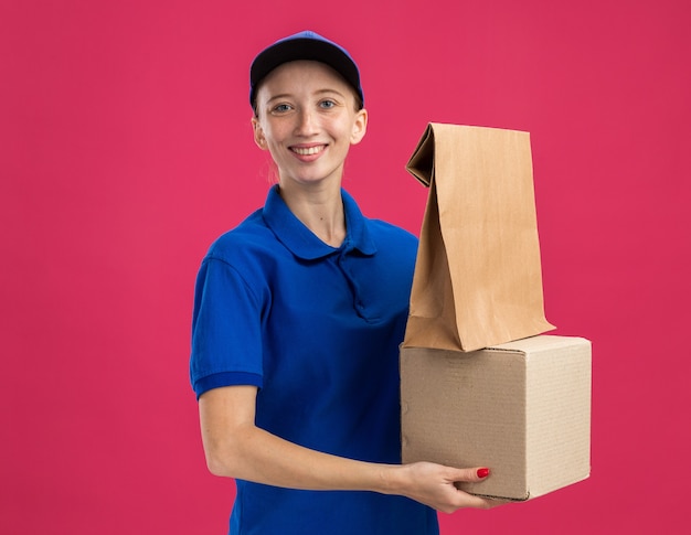 Repartidor joven en uniforme azul y gorra con caja de cartón y paquete de papel mirando camrera sonriendo confiado de pie sobre la pared rosa