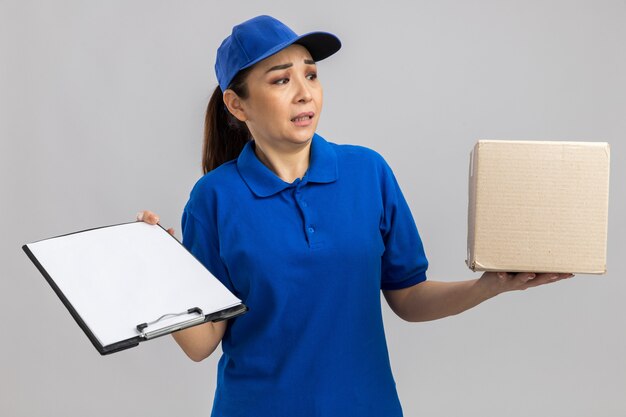 Repartidor joven en uniforme azul y gorra con caja de cartón y portapapeles mirando a un lado confundido parado sobre pared blanca