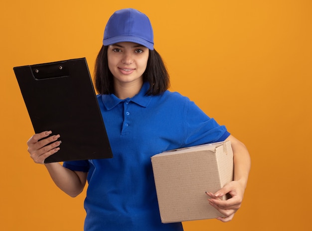 Repartidor joven en uniforme azul y gorra con caja de cartón y portapapeles sonriendo con cara feliz sonriendo de pie sobre la pared naranja