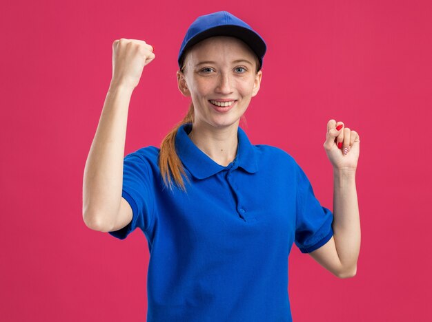 Repartidor joven en uniforme azul y gorra emocionados y felices apretando los puños de pie sobre la pared rosa