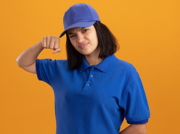 Foto gratuita repartidor joven en uniforme azul y gorra con expresión seria y segura mostrando puño parado sobre la pared naranja