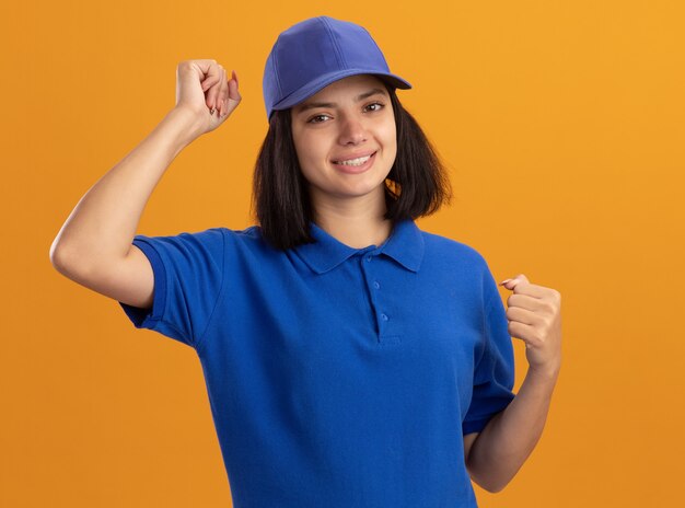 Repartidor joven en uniforme azul y gorra feliz y emocionado apretando los puños regocijándose de su éxito de pie sobre la pared naranja