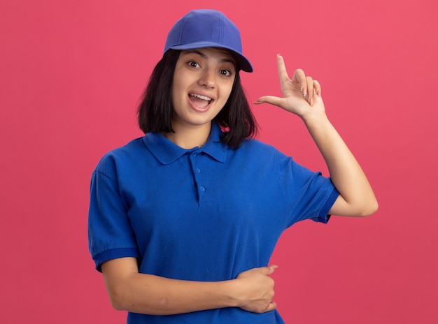 Repartidor joven en uniforme azul y gorra feliz y sorprendida mostrando el dedo índice teniendo una nueva idea de pie sobre la pared rosa