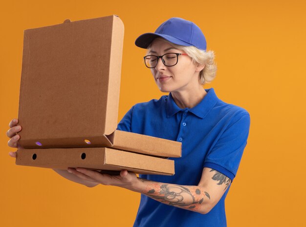 Repartidor joven en uniforme azul y gorra con gafas sosteniendo cajas de pizza abriendo una caja inhalando aroma agradable sobre pared naranja