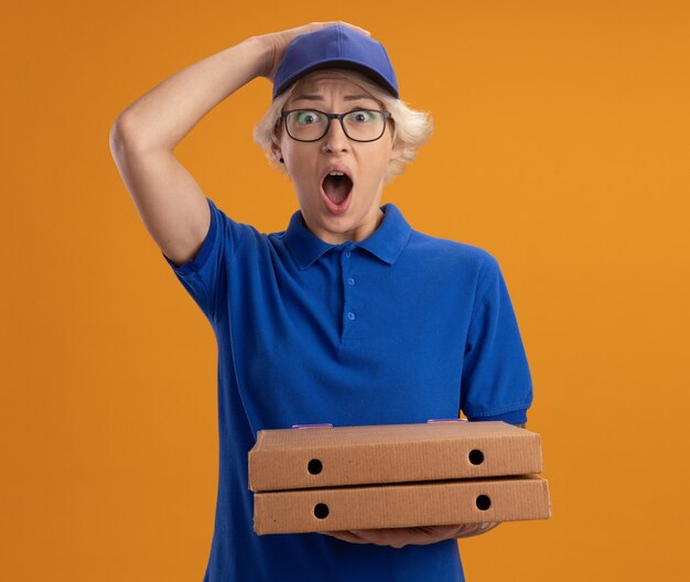 Repartidor joven en uniforme azul y gorra con gafas sosteniendo cajas de pizza asombrado y sorprendido por la pared naranja