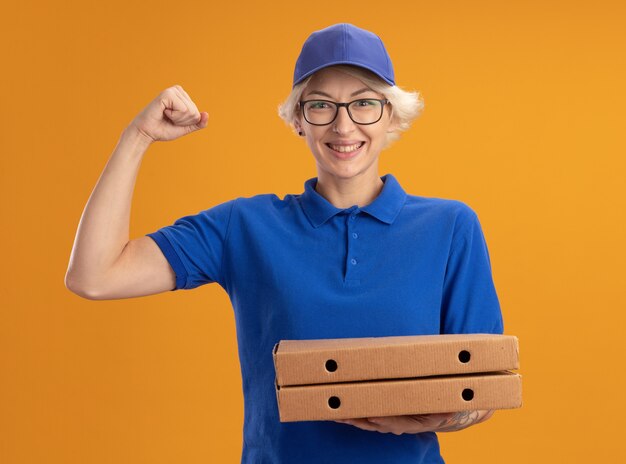Repartidor joven en uniforme azul y gorra con gafas sosteniendo cajas de pizza feliz y confiado apretando el puño sobre la pared naranja