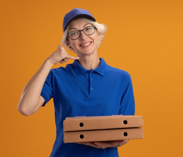 Repartidor joven en uniforme azul y gorra con gafas sosteniendo cajas de pizza sonriendo haciendo cal me gesto sonriendo sobre pared naranja