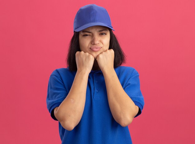 Repartidor joven en uniforme azul y gorra haciendo la boca torcida con expresión decepcionada de pie sobre la pared rosa