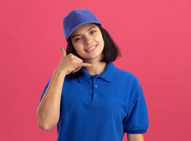 Repartidor joven en uniforme azul y gorra haciendo gesto de llamarme sonriendo de pie sobre la pared rosa