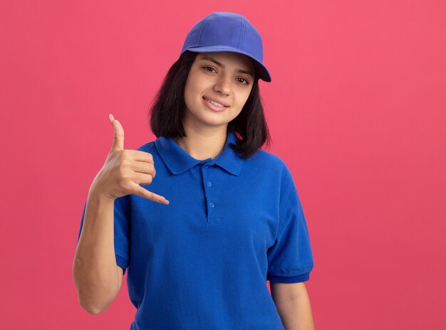 Repartidor joven en uniforme azul y gorra haciendo gesto de llamarme sonriendo de pie sobre la pared rosa