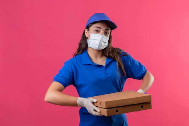 Repartidor joven en uniforme azul y gorra con máscara protectora facial y guantes sosteniendo cajas de pizza mirando a la cámara con expresión facial seria y segura de pie sobre fondo rosa