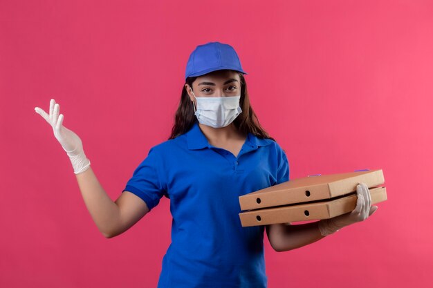 Repartidor joven en uniforme azul y gorra con máscara protectora facial y guantes sosteniendo cajas de pizza mirando a la cámara sonriendo seguro positivo y feliz de pie sobre fondo rosa