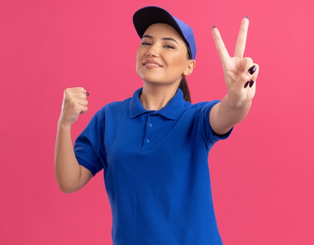 Foto gratuita repartidor joven en uniforme azul y gorra mirando al frente apretando el puño mostrando v-sign sonriendo alegremente de pie sobre la pared rosa